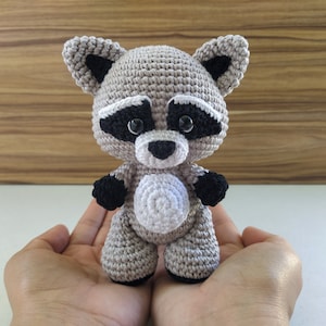 May include: A crocheted raccoon plush toy with black eyes, a black nose, and black and white markings on its face. The toy is standing on a person's hands.