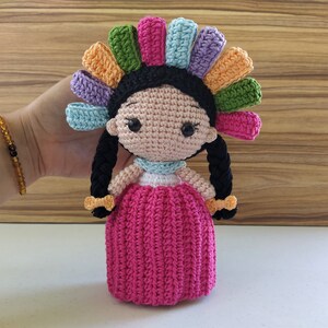 May include: A crocheted doll with black hair, a pink dress, and a colorful headdress. The doll is handmade and has a sweet, whimsical expression.