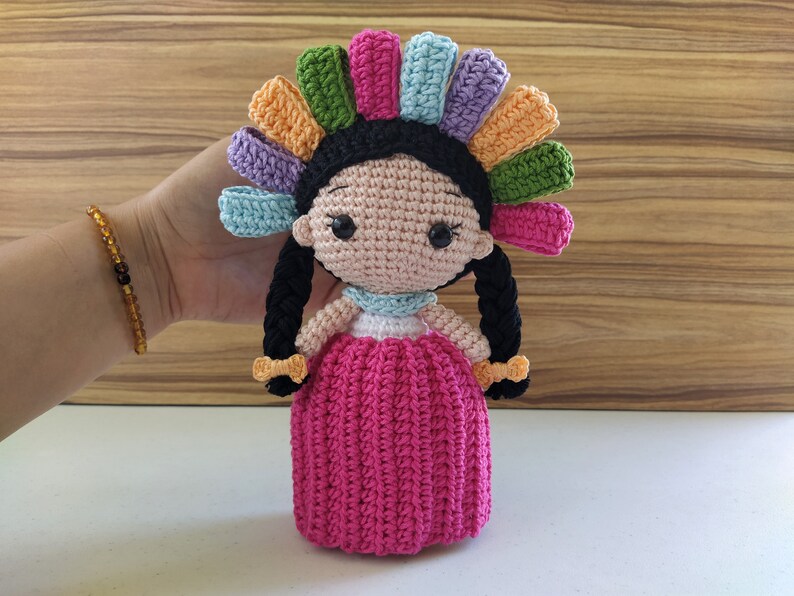 May include: A crocheted doll with black hair, a pink dress, and a colorful headdress. The doll is handmade and has a sweet, whimsical expression.