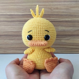 Puede incluir: Un pato amarillo de crochet con patas naranjas y ojos negros. El pato está sentado en las palmas de las manos de una persona.