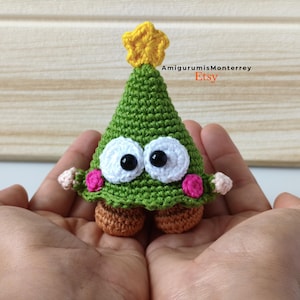 Puede incluir: Un árbol de Navidad verde tejido a mano con grandes ojos blancos, pupilas negras y mejillas rosas. El árbol tiene una estrella amarilla en la parte superior y pies marrones. El nombre de la tienda Etsy "AmigurumisMonterrey" es visible.