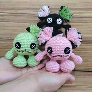 May include: Three handmade crochet dolls in green, pink, and black, each with a round head, stitched eyes, and a stitched mouth. The dolls have small arms and legs, and tufts of yarn for hair. The dolls are held in a person's hands.