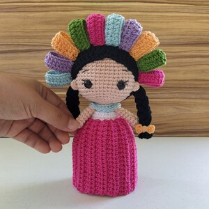 May include: A crocheted doll with a colorful flower crown, pink dress, and black hair. The doll is handmade and has a sweet, whimsical expression.