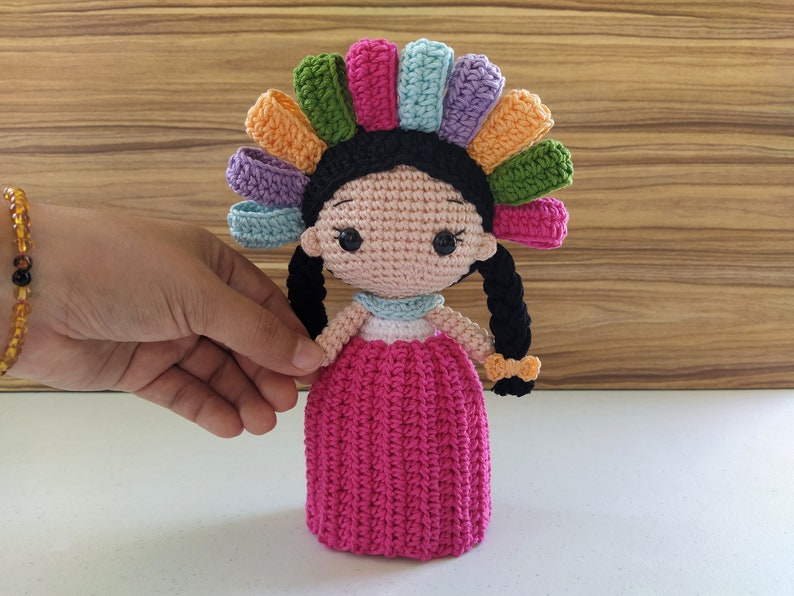 May include: A crocheted doll with a colorful flower crown, pink dress, and black hair. The doll is handmade and has a sweet, whimsical expression.