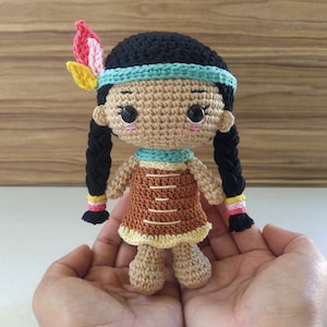 Puede incluir: Muñeca de crochet con cabello castaño, un vestido marrón y amarillo y una diadema azul. La muñeca lleva un tocado de plumas con plumas rosas, amarillas y naranjas.