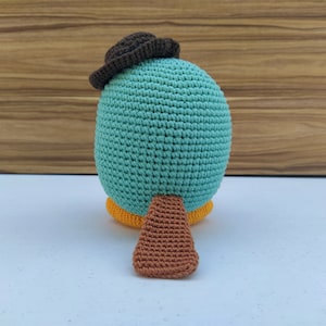 May include: A crocheted plush toy shaped like a platypus wearing a brown hat. The toy is teal blue with a brown tail and yellow feet.