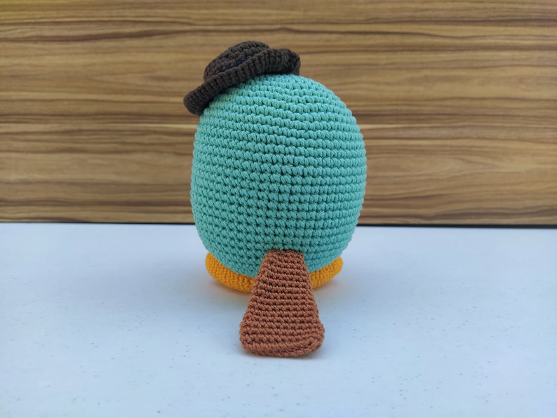 May include: A crocheted plush toy shaped like a platypus wearing a brown hat. The toy is teal blue with a brown tail and yellow feet.