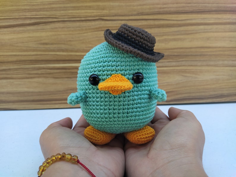 May include: A crocheted teal blue platypus wearing a brown cowboy hat with orange feet. The platypus is sitting in the palms of two hands.
