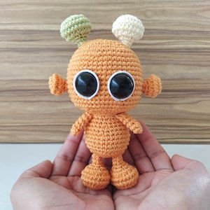 May include: A small orange crocheted alien toy with large black eyes and two antennas, one green and one white, is held in the palms of two hands.