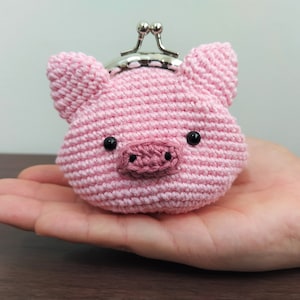 Peut inclure: Une petite bourse à monnaie en forme de cochon en crochet rose avec des yeux noirs et un groin rose. La bourse a un fermoir argenté.