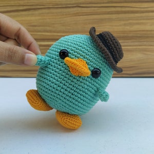 May include: A teal blue crocheted bird wearing a brown hat with black eyes and an orange beak. The bird has orange feet and is standing on a white surface.