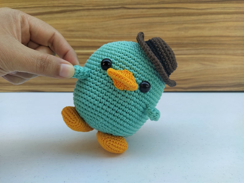 May include: A teal blue crocheted bird wearing a brown hat with black eyes and an orange beak. The bird has orange feet and is standing on a white surface.