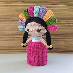 May include: A crocheted doll with black hair, a pink dress, and a colorful flower crown.