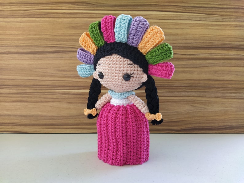 May include: A crocheted doll with black hair, a pink dress, and a colorful flower crown.