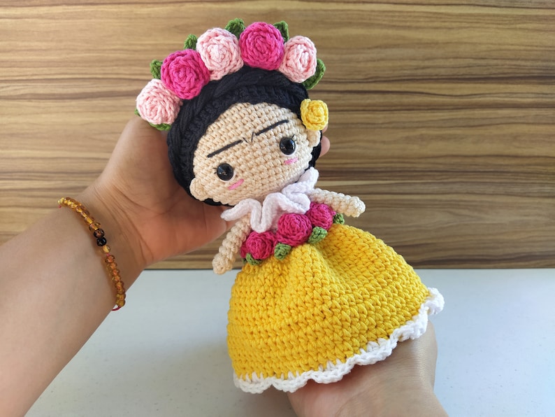 May include: Crocheted doll with a yellow dress, white lace trim, and a pink flower crown. The doll has black hair and a painted face.