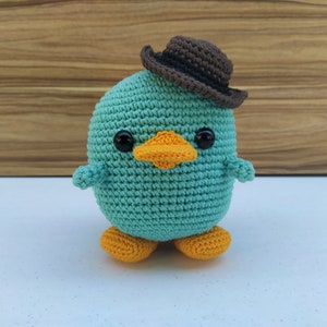 May include: A teal crocheted plush toy shaped like a duck wearing a brown felt cowboy hat. The duck has orange feet and black eyes.