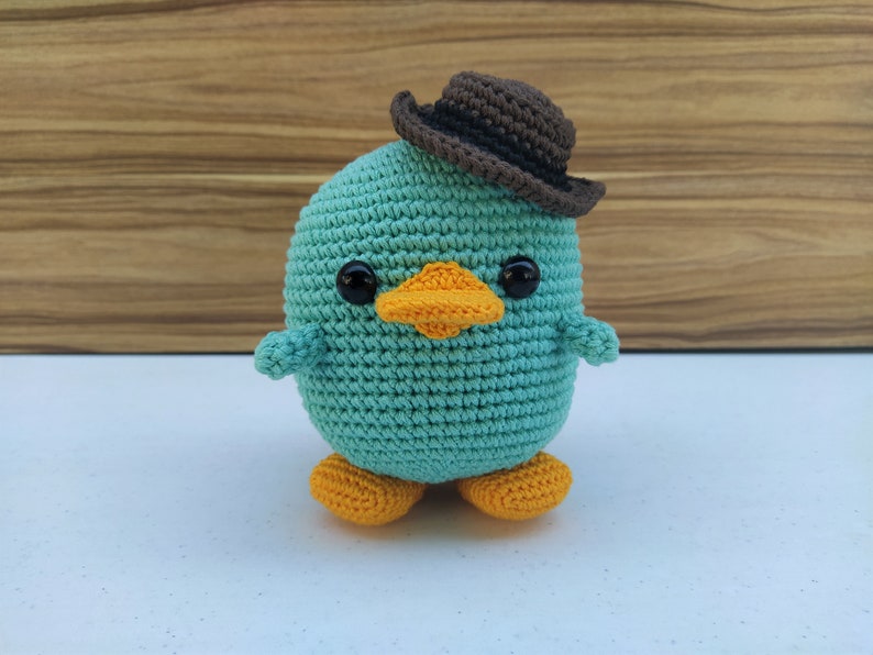 May include: A teal crocheted plush toy shaped like a duck wearing a brown felt cowboy hat. The duck has orange feet and black eyes.