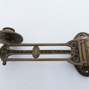 May include: Antique brass wall-mounted candle holder with ornate details and a swiveling arm. The holder has a round base with a decorative border and a hinged arm that can be adjusted to different angles.