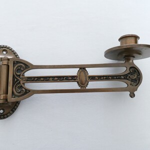 May include: A vintage brass wall-mounted candle holder with an ornate design. The holder has a hinged arm that can be adjusted to different angles. The candle holder is in good condition with some signs of age.