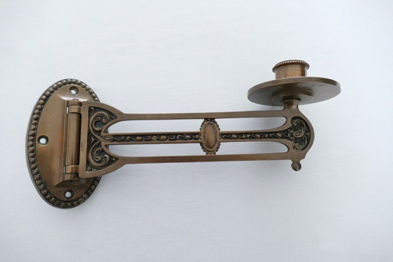 May include: A vintage brass wall-mounted candle holder with an ornate design. The holder has a hinged arm that can be adjusted to different angles. The candle holder is in good condition with some signs of age.