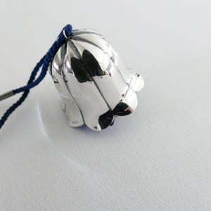 Op de afbeelding: Een zilveren, klokvormig ornament met een donkerblauwe koord. De bel heeft een reflecterend oppervlak en een bloemenontwerp. Het koord is bevestigd aan de bovenkant van de bel, wat suggereert dat het kan worden opgehangen. De achtergrond is een effen wit oppervlak.