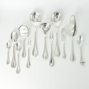 May include: A collection of eleven polished silver-toned metal utensils including various spoons, a ladle, a cake server, a butter knife, and two-pronged forks. The set features smooth, rounded handles and reflective surfaces arranged on a white background.
