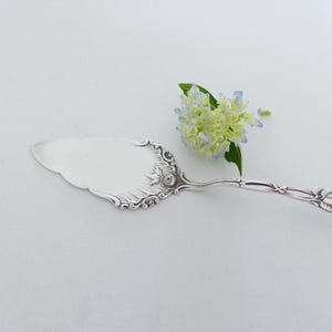 Op de afbeelding: Een zilveren taartschep met een sierlijk ontwerp met een roos aan het einde van de steel. De schep ligt op een witte achtergrond met een klein boeket lichtblauwe en witte bloemen.