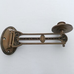 May include: A brass wall-mounted sconce with an ornate design. The sconce has a hinged arm and a round plate for holding a candle or other light source.
