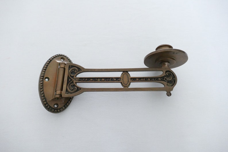 May include: A brass wall-mounted sconce with an ornate design. The sconce has a hinged arm and a round plate for holding a candle or other light source.