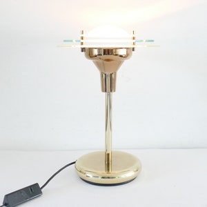 Op de afbeelding: Een goudkleurige tafellamp met een witte glazen kap en een ronde voet. De lamp heeft een modern ontwerp met een strakke, minimalistische esthetiek.