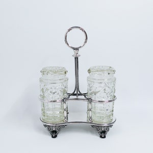 Op de afbeelding: Zilveren geplateerd condimentenset met twee heldere glazen potten met een geslepen glasontwerp. De set heeft een decoratief handvat en staat op een voet met drie poten.