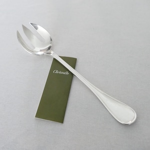 May include: A silver serving fork with three tines, resting on a green card with the brand name "Christofle" printed on it. The fork has a long, slender handle with a rounded end. The background is a light gray surface.