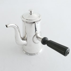 Puede incluir: Una cafetera plateada pulida con un asa negra. La cafetera tiene un pico curvo y una tapa con un pequeño pomo. El asa es rectangular y está unida al lateral. Diseño clásico.