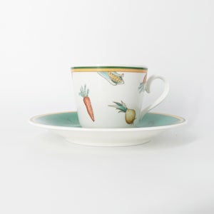 Puede incluir: Un juego de taza de té y platillo de cerámica blanca con un patrón de vegetales coloridos que incluye zanahorias, cebollas y maíz. La taza tiene un asa curva y un borde con bandas finas verdes y amarillas, y el platillo tiene un borde ancho color verde azulado.