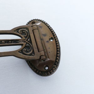 May include: A brass guitar strap button with an ornate design. The button has a hinge and is oval shaped.