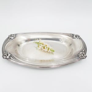Könnte beinhalten: Ein ovales Tablett aus silberfarbenem Metall mit einer glatten, reflektierenden Oberfläche und erhabenen Blumenmotiven an allen vier Ecken. Ein kleiner Zweig mit winzigen weißen Blüten und grünen Stielen liegt in der Mitte der flachen Schale.