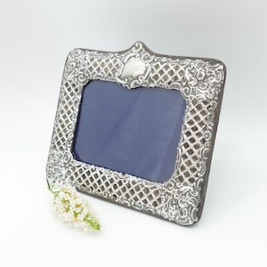 May include: A rectangular silver-toned picture frame featuring an intricate lattice pattern and ornate scrollwork borders. The frame has a central dark blue insert and a small white floral sprig positioned at the bottom left corner of the frame.