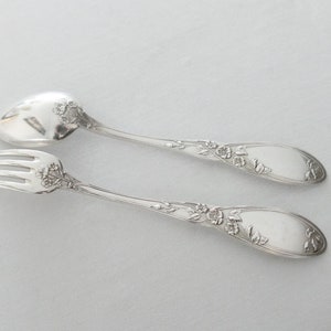 Puede incluir: Un juego de cuchara y tenedor de servir plateados. Los cubiertos tienen un diseño floral en los mangos y cerca de las cabezas de los utensilios. La cuchara tiene un cuenco ranurado. El juego está sobre un fondo blanco.