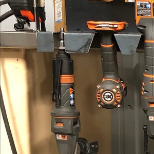 May include: A close-up of a black and orange power tool hanging on a metal rack. The tool has a black handle and a silver blade. The tool is hanging on a metal rack with a black background.