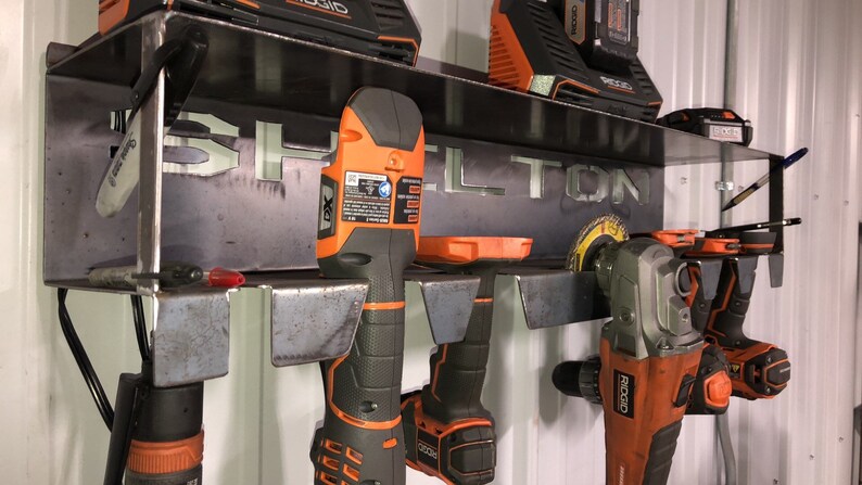 May include: A metal wall-mounted tool rack with the name "SHELTON" cut out. The rack holds a variety of power tools, including drills, grinders, and a saw. The tools are all orange and black.