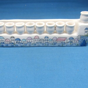 May include: White ceramic menorah with a blue and white painted design of a cityscape. The menorah has nine candle holders and a central spout.