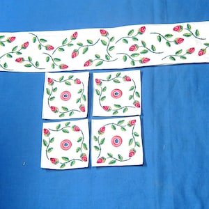 May include: A white table runner and four matching square coasters. Each item features a repeating pattern of pink roses, green leaves, and stems. The items are arranged on a blue surface.