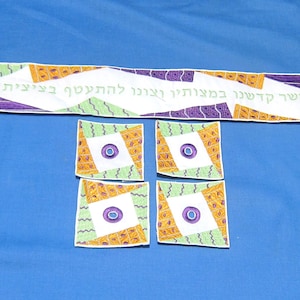 May include: A white textile set with a long banner and four square coasters. The banner has text and geometric patterns in purple, orange, and green. The coasters feature a central circle design.