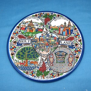 May include: A round ceramic plate with a blue border and a white center. The plate is decorated with colorful images of Jerusalem, Bethlehem, Nazareth, and the Holy Land. The plate is a souvenir from Israel.