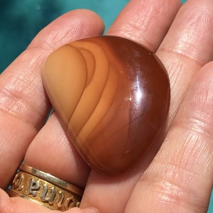Pode incluir: Uma pedra de cornalina lisa e polida, em forma de cora&ccedil;&atilde;o, de cor marrom avermelhada com um padr&atilde;o em camadas de tons de marrom mais claros e mais escuros.