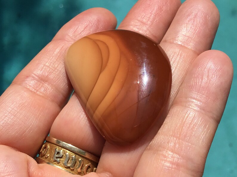 Pode incluir: Uma pedra de cornalina lisa e polida, em forma de cora&ccedil;&atilde;o, de cor marrom avermelhada com um padr&atilde;o em camadas de tons de marrom mais claros e mais escuros.