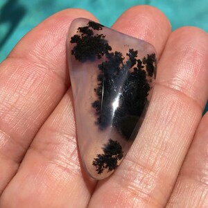 May include: A polished, translucent, light grey agate with black dendritic patterns. The agate is shaped like a teardrop and has a smooth, polished surface.