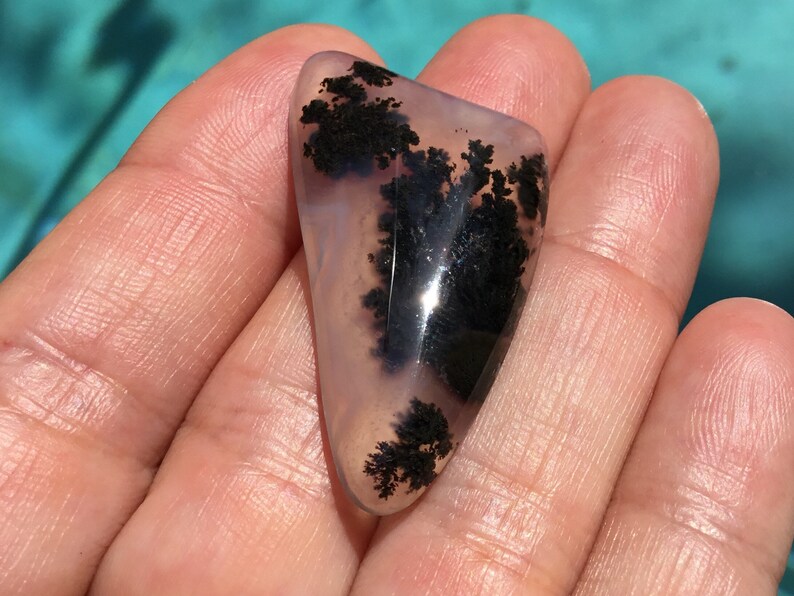 May include: A polished, translucent, light grey agate with black dendritic patterns. The agate is shaped like a teardrop and has a smooth, polished surface.