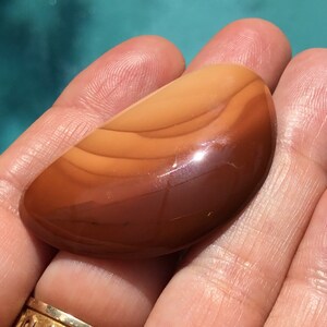 Pode incluir: Um caboch&atilde;o de &aacute;gata marrom polido e liso com bandas laranja e brancas. A pedra tem formato oval e um acabamento brilhante.