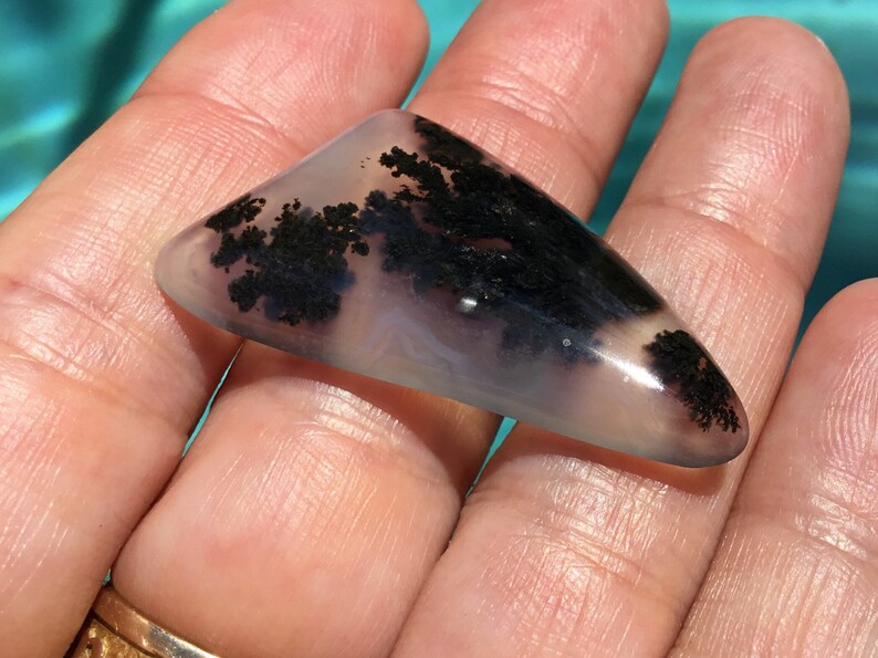 May include: A triangular, translucent, light grey agate cabochon with black dendritic inclusions. The agate has a smooth, polished surface.